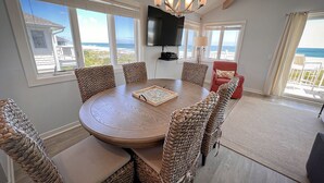 Une table ronde en bois, entourée de chaises en osier, un téléviseur à écran plat fixé au mur et une vue sur l’océan par de grandes fenêtres.