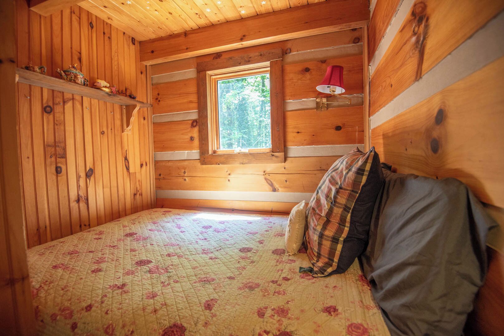 A cozy cabin room with a wooden interior, a single bed with floral bedding, a window with a view of trees, and a small shelf with decorative items.