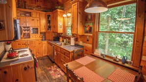 A cabin-style kitchen with wooden cabinets, a stove, and a large window overlooking a forest. - Creekside Escape | Hot Tub & Wooded Privacy | Prime West Jefferson Location (West Jefferson)