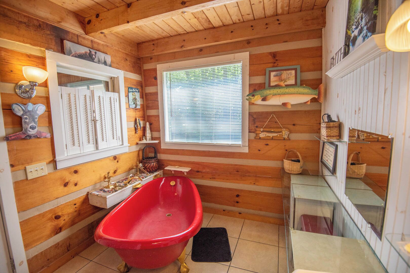 A cabin-style bathroom with a red bathtub, wooden walls, a window with blinds, and various decorations including a fish, a deer head, and framed pictures.
