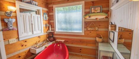 A cabin-style bathroom with a red bathtub, wooden walls, a window with blinds, and various decorations including a fish, a deer head, and framed pictures.