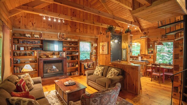 A cozy log cabin interior with a fireplace, wooden shelves, a television, and a dining area. - Creekside Escape | Hot Tub & Wooded Privacy | Prime West Jefferson Location (West Jefferson)