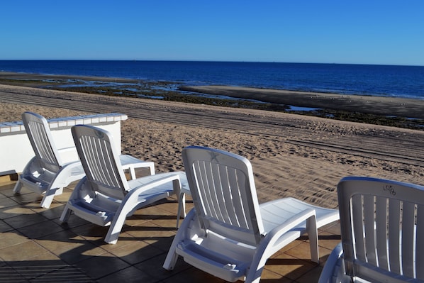 Un espacio de descanso frente a la playa, con sillas reclinables blancas y una terraza con baldosas.