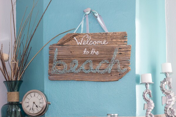 A wooden sign with "Welcome to the beach" written on it, a clock, and decorative items on a teal wall.