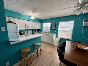A compact kitchen with teal walls, white cabinets, a microwave, stove, and a ceiling fan. There are two stools and a dining table with a fruit bowl.