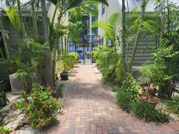 A paved pathway leading to a gated entrance, surrounded by lush greenery and potted plants.