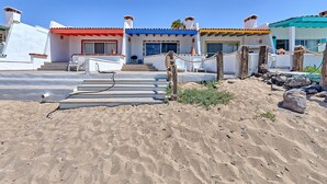 A beachfront property with a series of white buildings, each with a distinct colored roof. There are steps leading to the entrance, and a rope barrier separates the beach from the property. - Buenos Tiempos – Beachfront, Sleeps 6, Rocky Point (Puerto Peñasco)