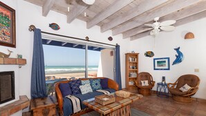 A living room with a view of the beach, a ceiling fan, and a bookshelf. - Buenos Tiempos – Beachfront, Sleeps 6, Rocky Point (Puerto Peñasco)