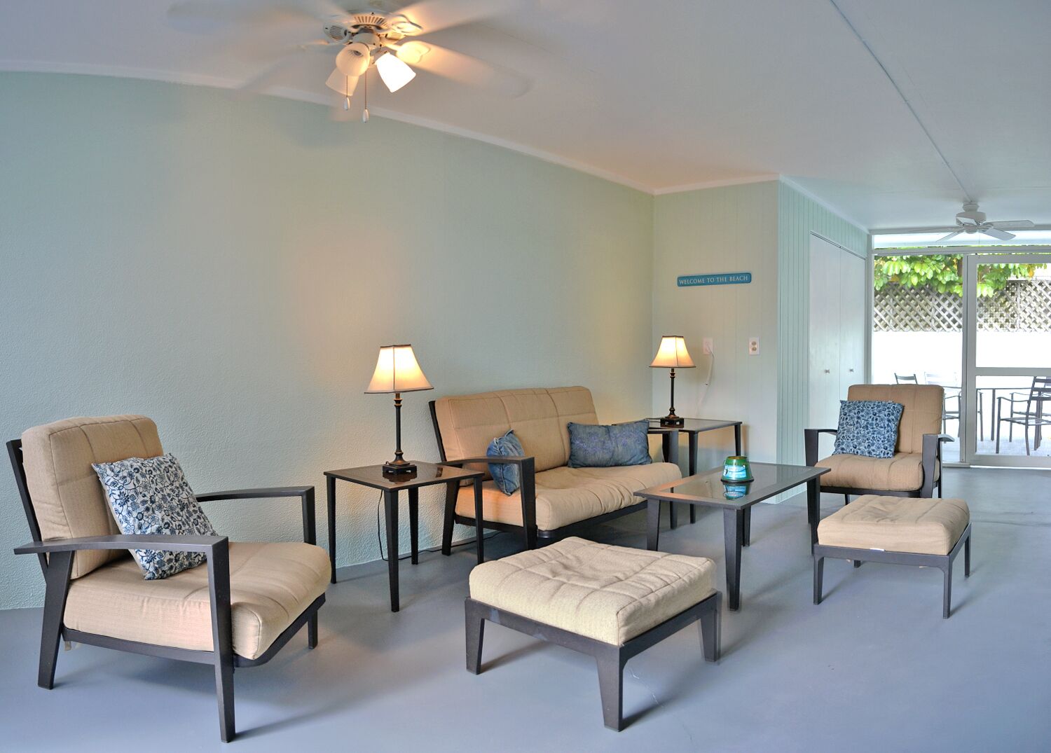 A modern living room with a sofa, two armchairs, a coffee table, and a side table. There is a ceiling fan and a sign that reads "Welcome to the Beach."