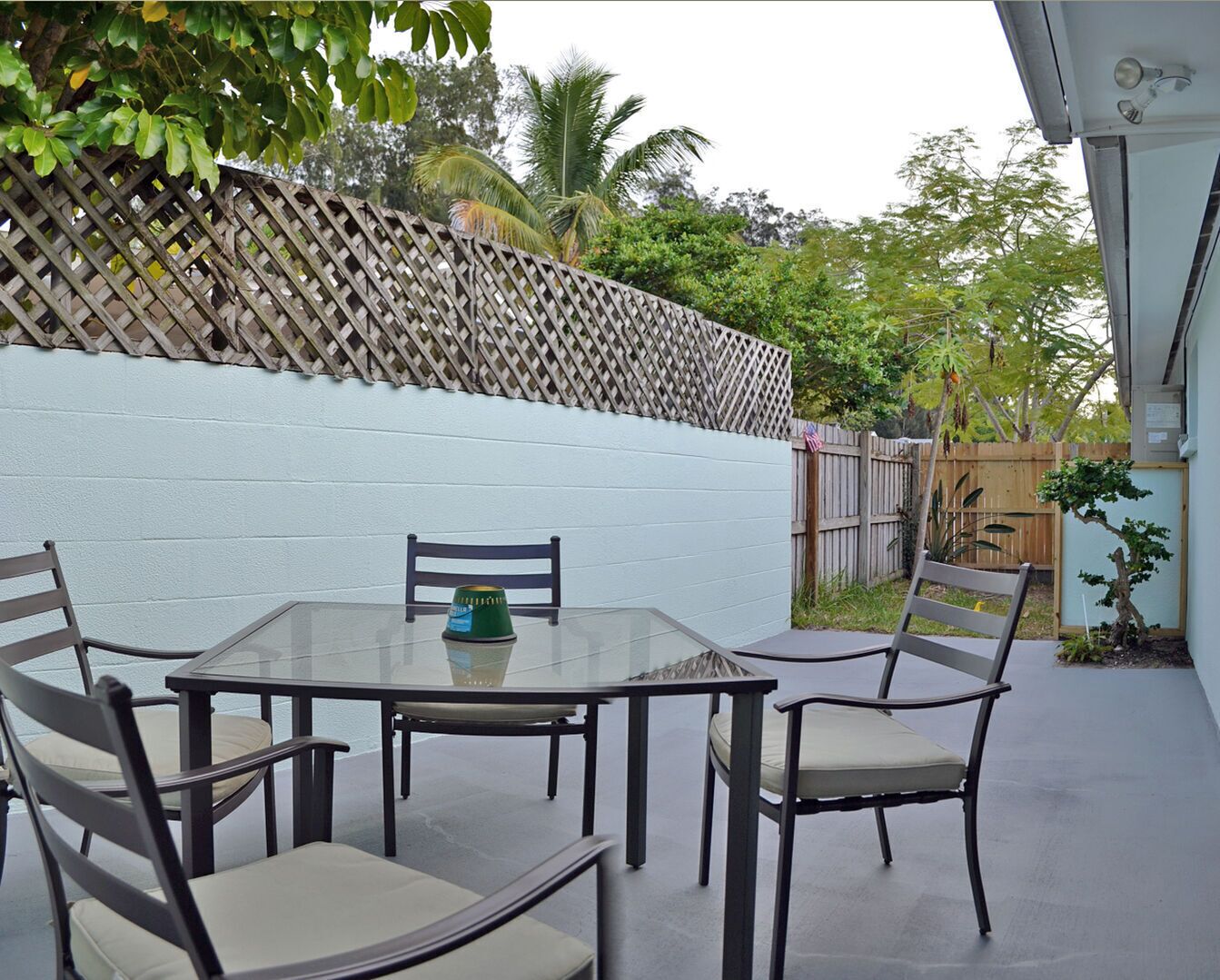 Un patio exterior con una mesa de vidrio y cuatro sillas, rodeado por un muro blanco y una valla de madera.