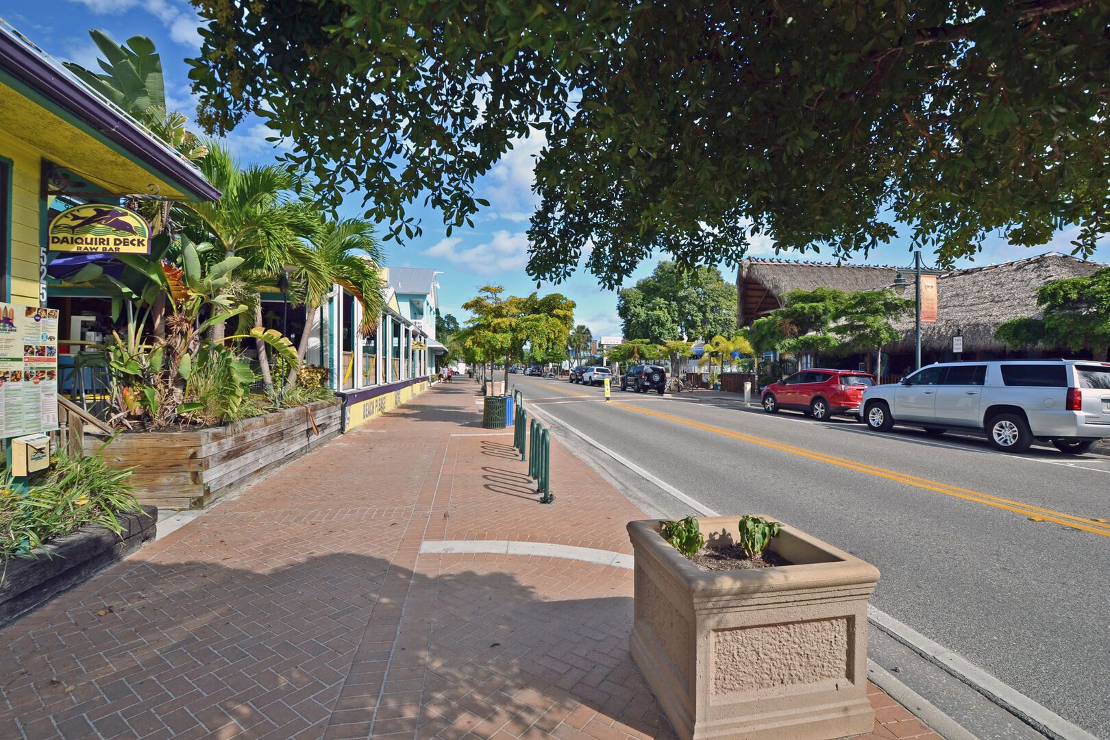 A street with a restaurant named 'Daquiri Deck' on the left, a sidewalk with planters, and a road with parked cars.