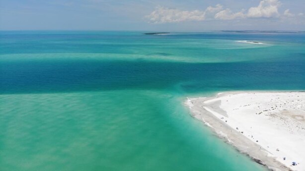 Un paysage côtier caractérisé par une eau turquoise cristalline, une plage de sable et une étendue terrestre visible au loin.