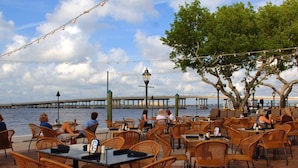 Un espace de restauration en plein air, avec des chaises et des tables en osier, offrant une vue sur un plan d’eau et un pont.