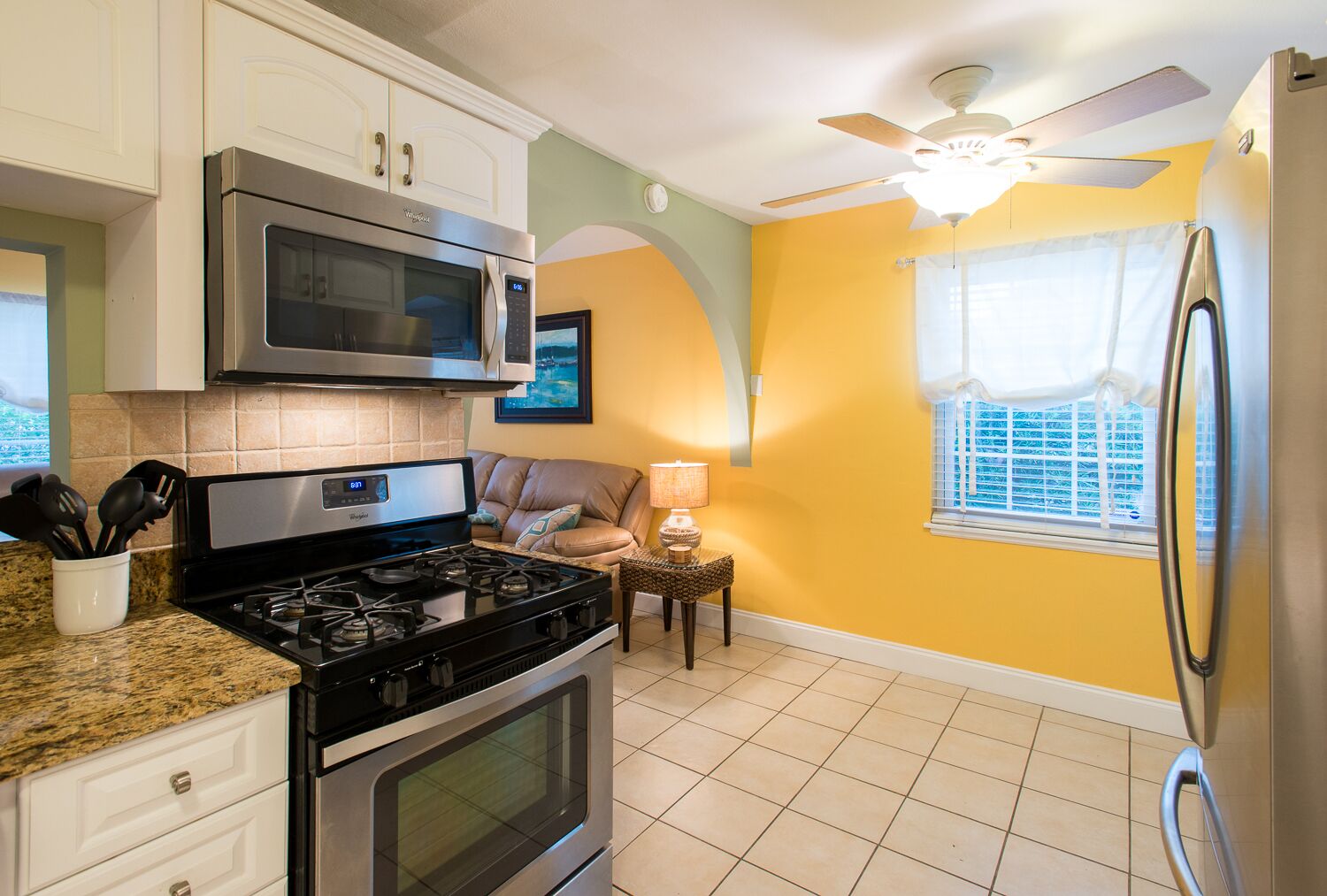 A kitchen with a gas stove, microwave, oven, and refrigerator. There is a ceiling fan, a window with blinds, and a small table with a lamp.