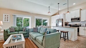 A modern living room with a green sectional sofa, a glass coffee table, and a kitchen area featuring white cabinetry, a stainless steel refrigerator, and a microwave. - Beach Days, Poolside Nights & Island Memories (Holmes Beach)