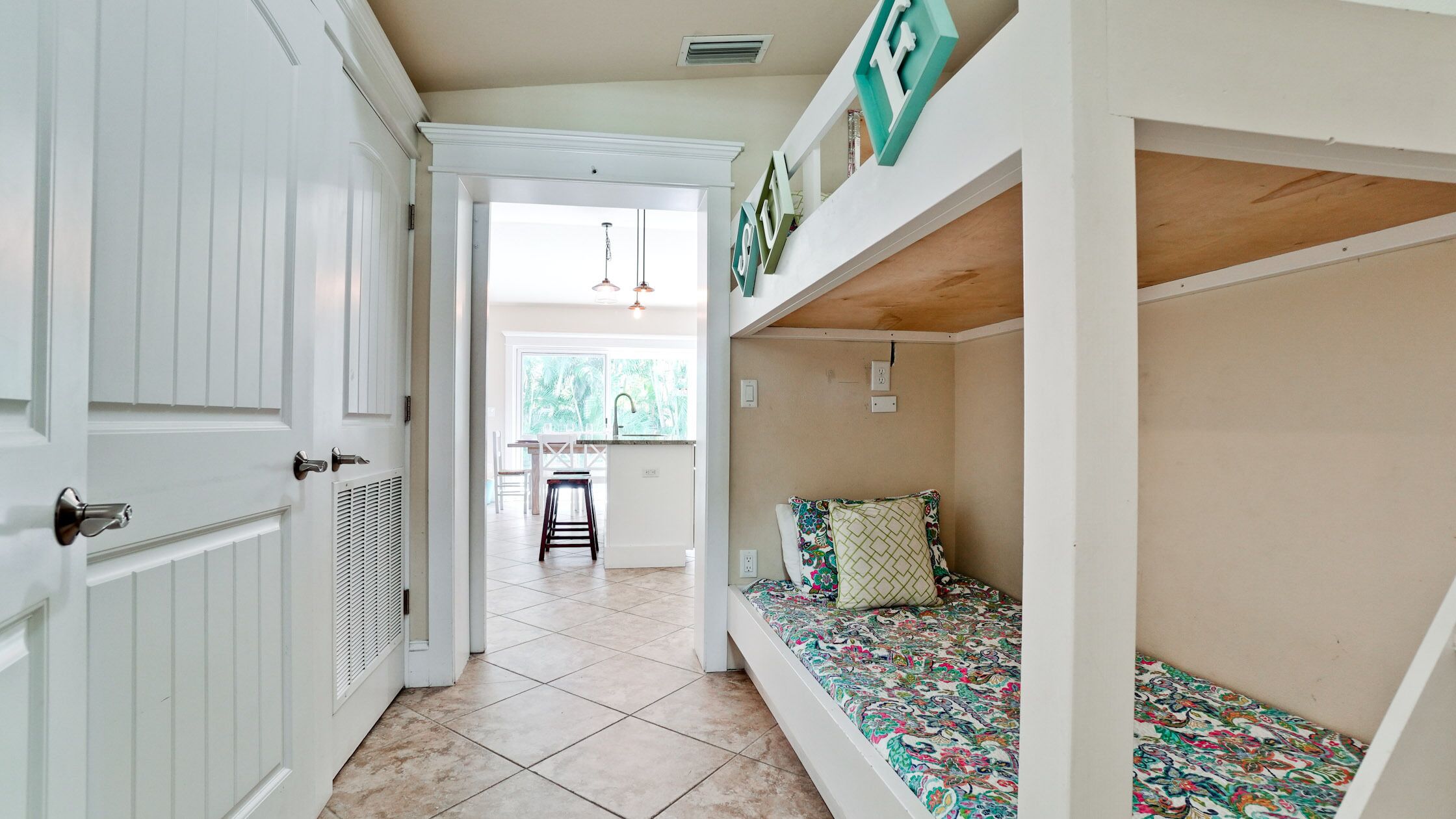A narrow hallway with a white door, a bench with colorful cushions, and a view into a kitchen area with a window and a hanging light fixture.
