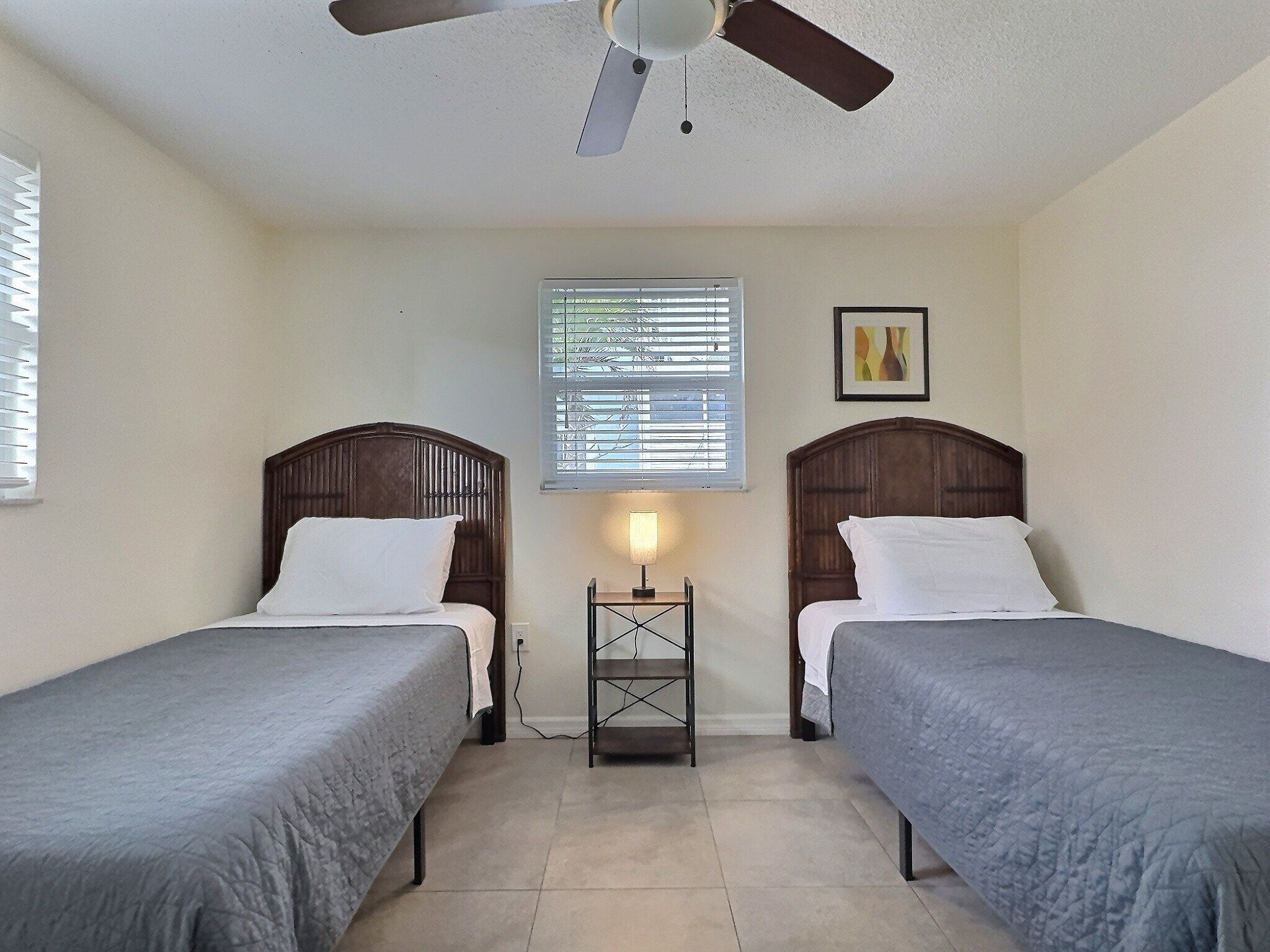 A room with two single beds, a ceiling fan, a window with blinds, a bedside table with a lamp, and a framed picture on the wall.