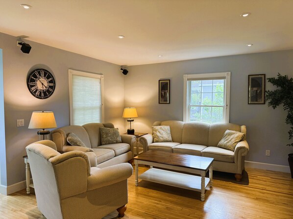 A living room with two couches, a coffee table, a wall clock, and a window with blinds.