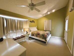 A bedroom with a bed, a chair, a table, a ceiling fan, and a window with curtains.