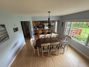 A dining area with a wooden table and chairs, a kitchen in the background, and a view of a red fence through a window.