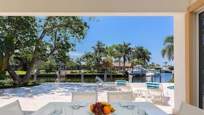 Una mesa de comedor con un plato de fruta, sillas transparentes y vista a un puerto deportivo con barcos y palmeras.
