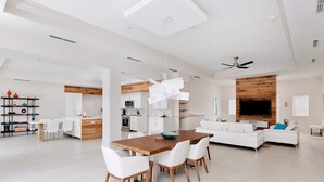 Amplia sala de estar con mesa de comedor de madera, sillas blancas, sofá blanco y televisor montado en una pared de madera.