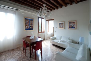 A living room with a white sofa, a wooden dining table with red chairs, a chandelier, and two framed artworks on the wall.