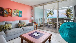 A living room with a sofa, a coffee table, and a teal bean bag. There are paintings on the wall and a view of the ocean through the sliding glass door. - 205E La Brisa-2BR 2BA beachfront condo w/parking, shared pool, grills (Key West)