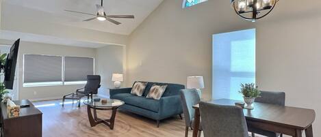 A spacious living room with a ceiling fan, a dining area with a table and chairs, a blue sofa, and a chandelier.