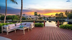 A wooden deck with a sofa and table, overlooking a calm body of water and a sunset. - 5 Bedroom Private Waterfront Villa with Pool and Dock (Fort Lauderdale)