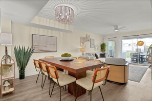 Un espace de repas moderne avec une table en bois, des chaises rembourrées, un lustre et une vue sur l’extérieur.