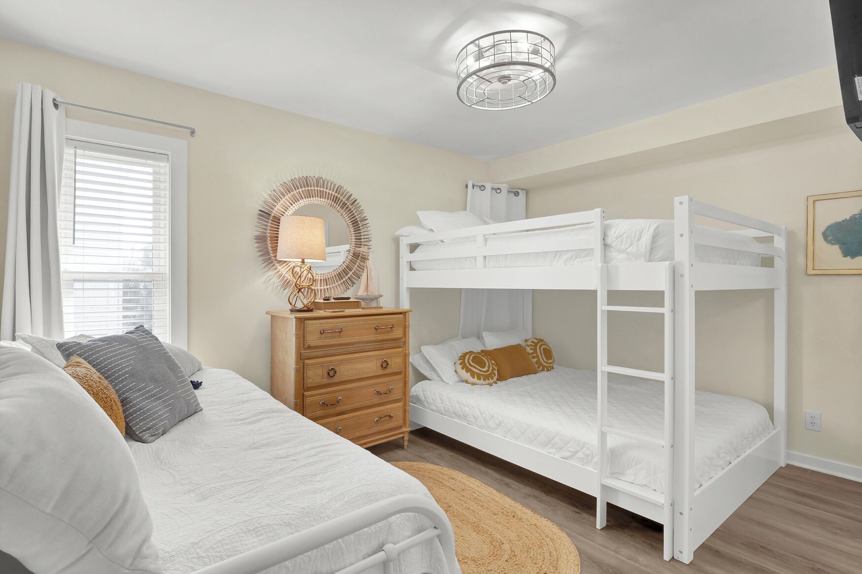 Une chambre avec un lit superposé, une table de chevet avec une lampe, une commode et une fenêtre avec des stores.