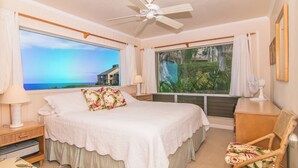 Un dormitorio con una cama grande, ventilador de techo, escritorio y dos ventanas con vistas al océano y un jardín.