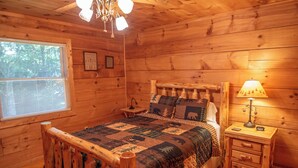 A wooden cabin bedroom with a bed, a nightstand, a lamp, and a window. - Nature Hideout with Great Location | Bear's Den Log Cabin | Hot Tub & Two Decks (Fleetwood)