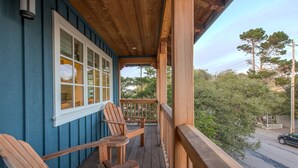 A wooden porch with two Adirondack chairs, a window with multiple panes, and a view of trees and a street. - 3684 Jewel by the Sea ~ Walk to Town and the Beach (Pacific Grove)
