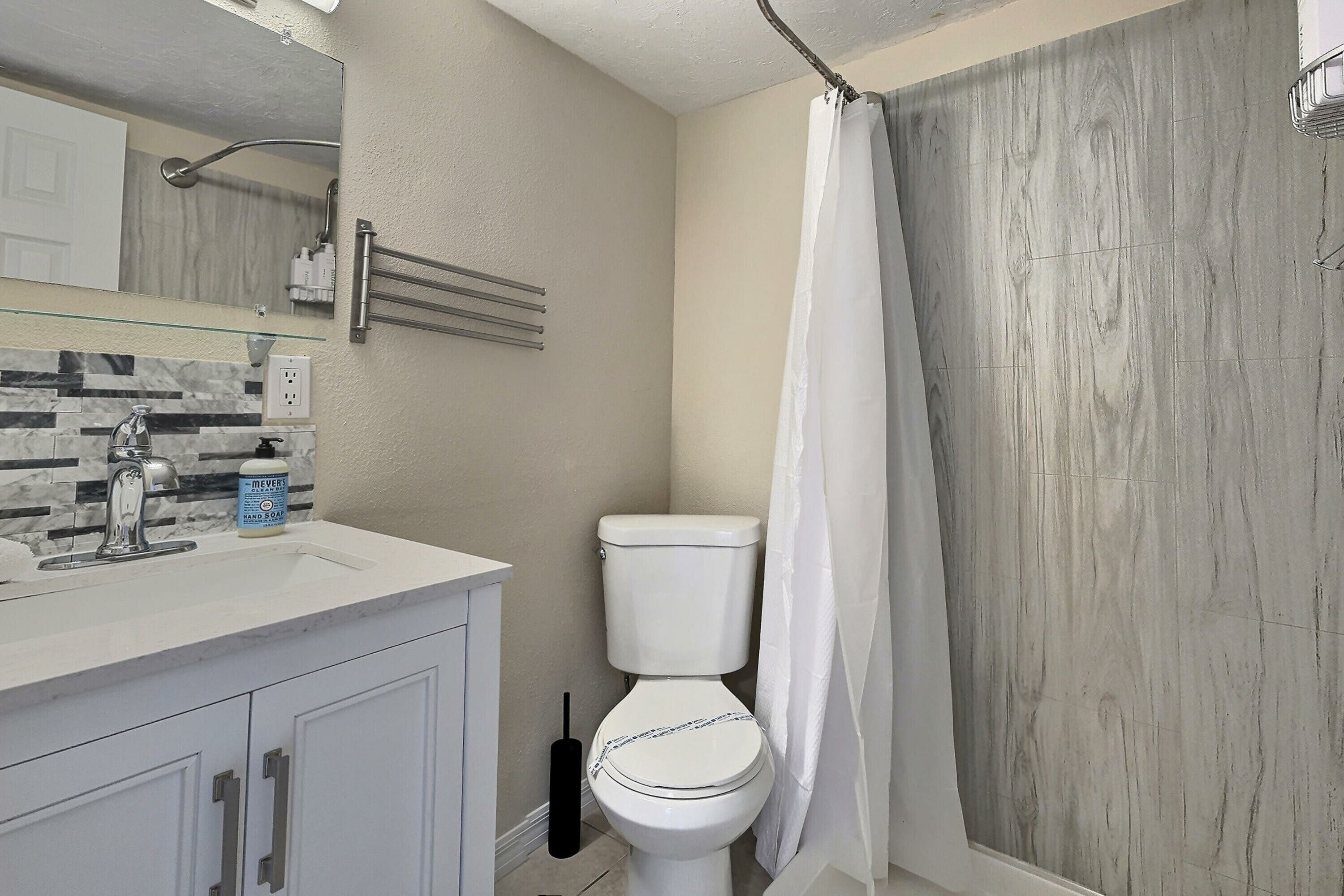 Uma casa de banho com sanita, lavatório com dispensador de sabonete, cortina de chuveiro e um porta-toalhas.