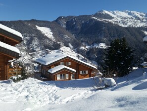 Een houten hut met een met sneeuw bedekte dak, omgeven door een besneeerd landschap en bergen op de achtergrond.