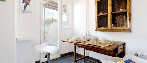 A small, clean room with a white sink, a wooden table with cups and a newspaper, a window with a mesh screen, and a framed bird picture on the wall.