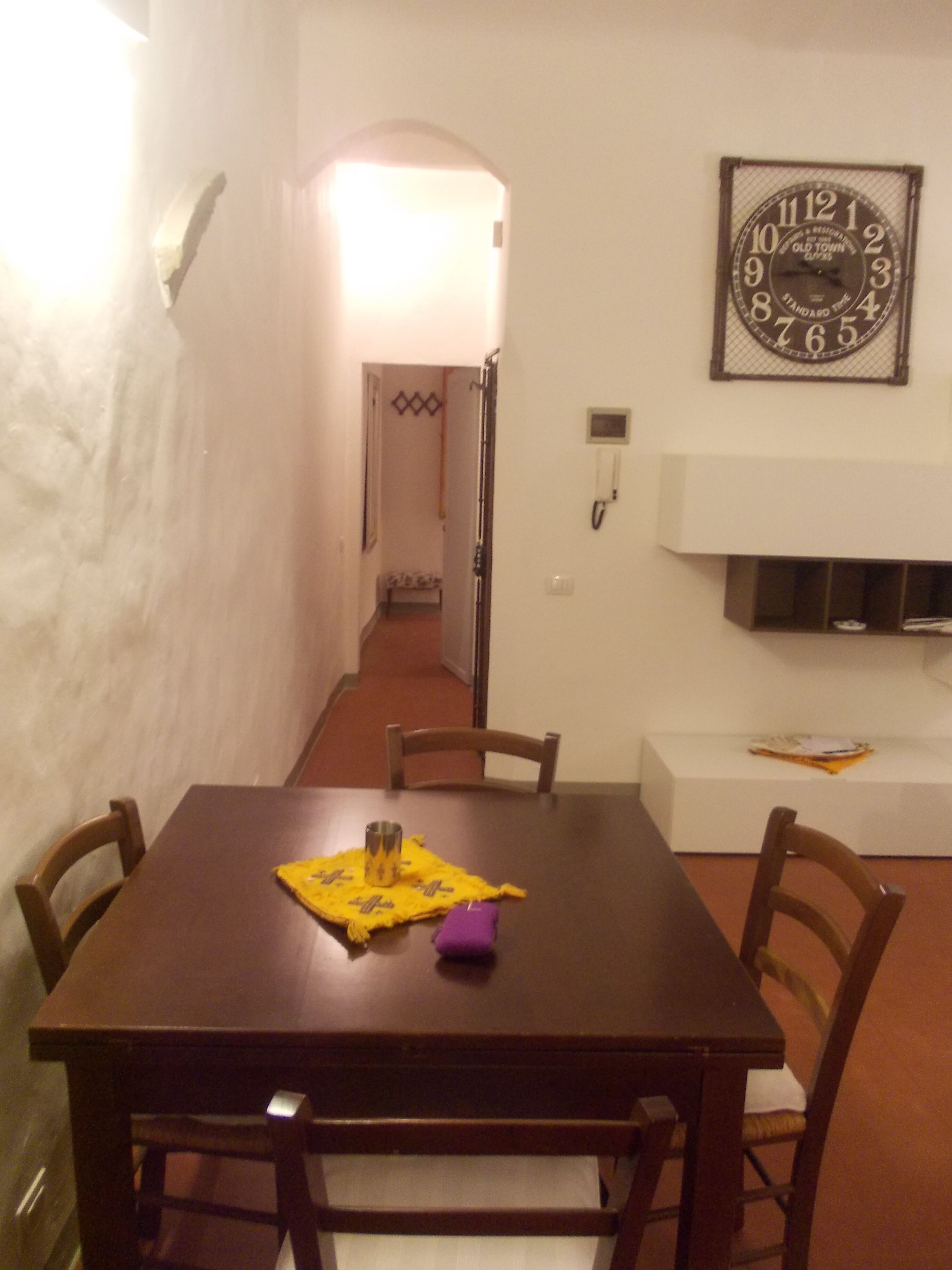 Una sala da pranzo con un tavolo di legno, quattro sedie, un sottopiatti giallo e un orologio appeso al muro.