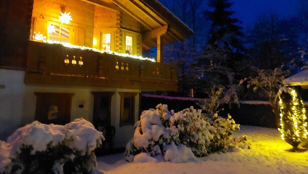 A wooden house with a balcony, snow-covered bushes, and a snow-covered roof.