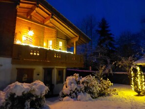 A wooden house with a balcony, snow-covered bushes, and a snow-covered roof.