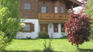A traditional wooden house with a balcony, surrounded by a well-maintained garden and a tree with autumn leaves.