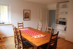 Un comedor con una mesa de madera, seis sillas, un camino de mesa rojo y blanco, un cuadro en la pared y un estante blanco con varios objetos.