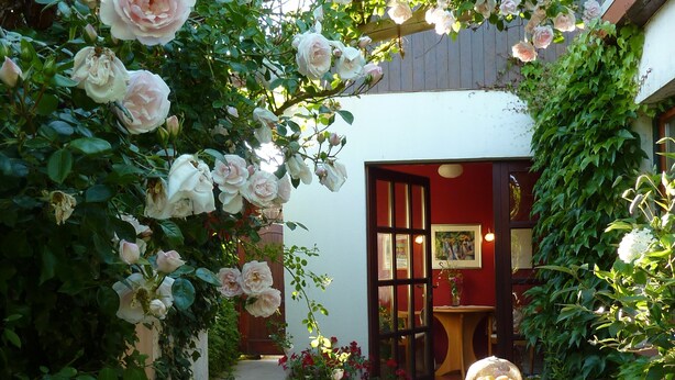 A garden with a wooden pergola covered in pink roses, a brick pathway, and a table with a lace tablecloth. - Garden idyll at the Wadden Sea National Park (Wesselburen)