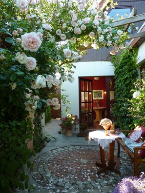 Un jardin agrémenté d’une pergola en bois recouverte de roses roses, d’un chemin en briques et d’une table recouverte d’une nappe en dentelle.