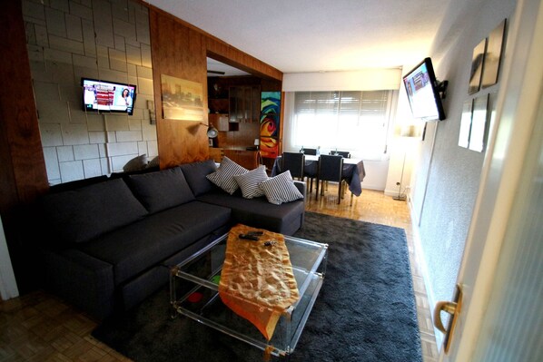 A modern living room with a sectional sofa, a glass coffee table, and a flat-screen TV mounted on the wall.