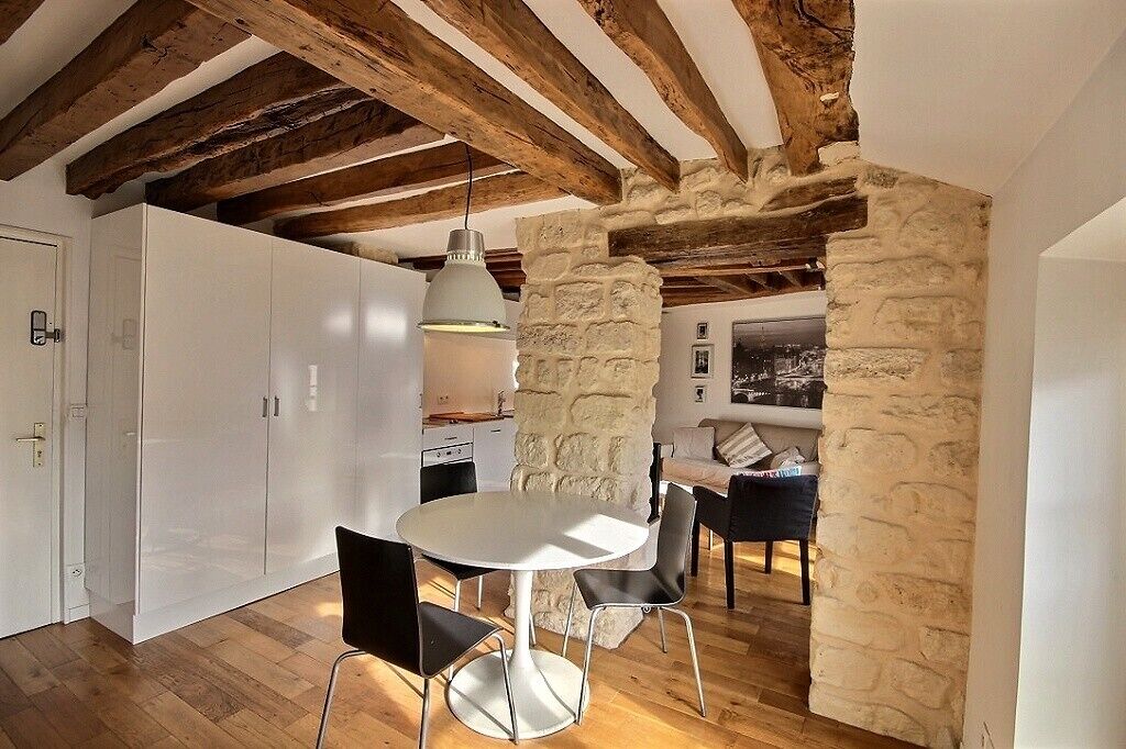 Une cuisine moderne avec des meubles blancs, une table de salle à manger ronde et des chaises noires. On y trouve un mur en pierre et des poutres en bois au plafond.