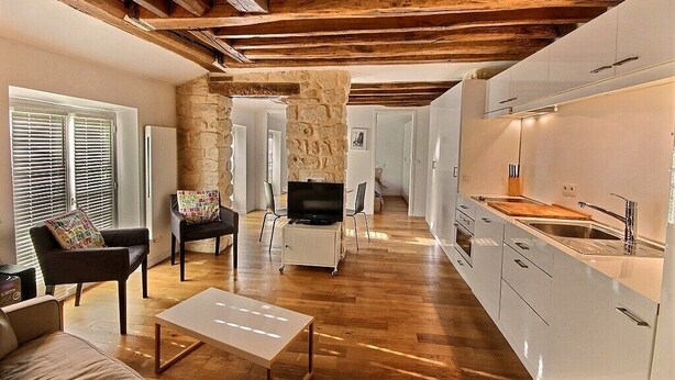 Un interior moderno con pared de piedra, vigas de madera, una cocina con armarios blancos y una zona de estar con televisor.