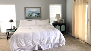 A bedroom with a bed, a ceiling fan, a painting, a bedside table with a lamp, and a window with blinds.