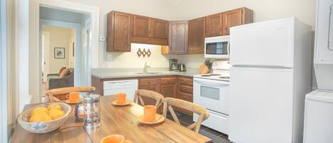 A kitchen with wooden cabinets, a white refrigerator, a microwave, and a dining table with orange cups and a bowl of oranges.